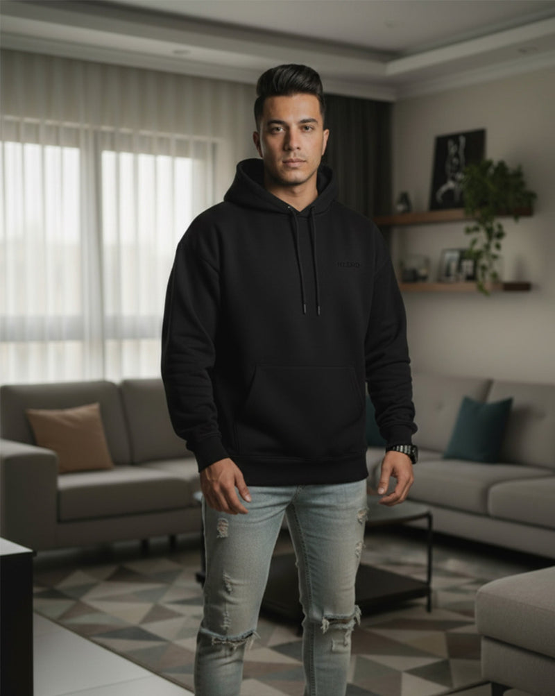 Man wearing a black hoodie and light blue jeans standing in a living room.