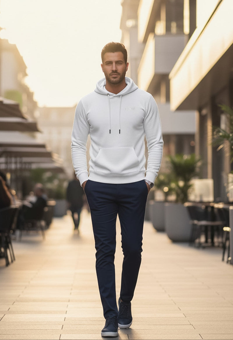 Man in a white hoodie and dark pants walking on a sidewalk with modern buildings and outdoor seating in the background.
